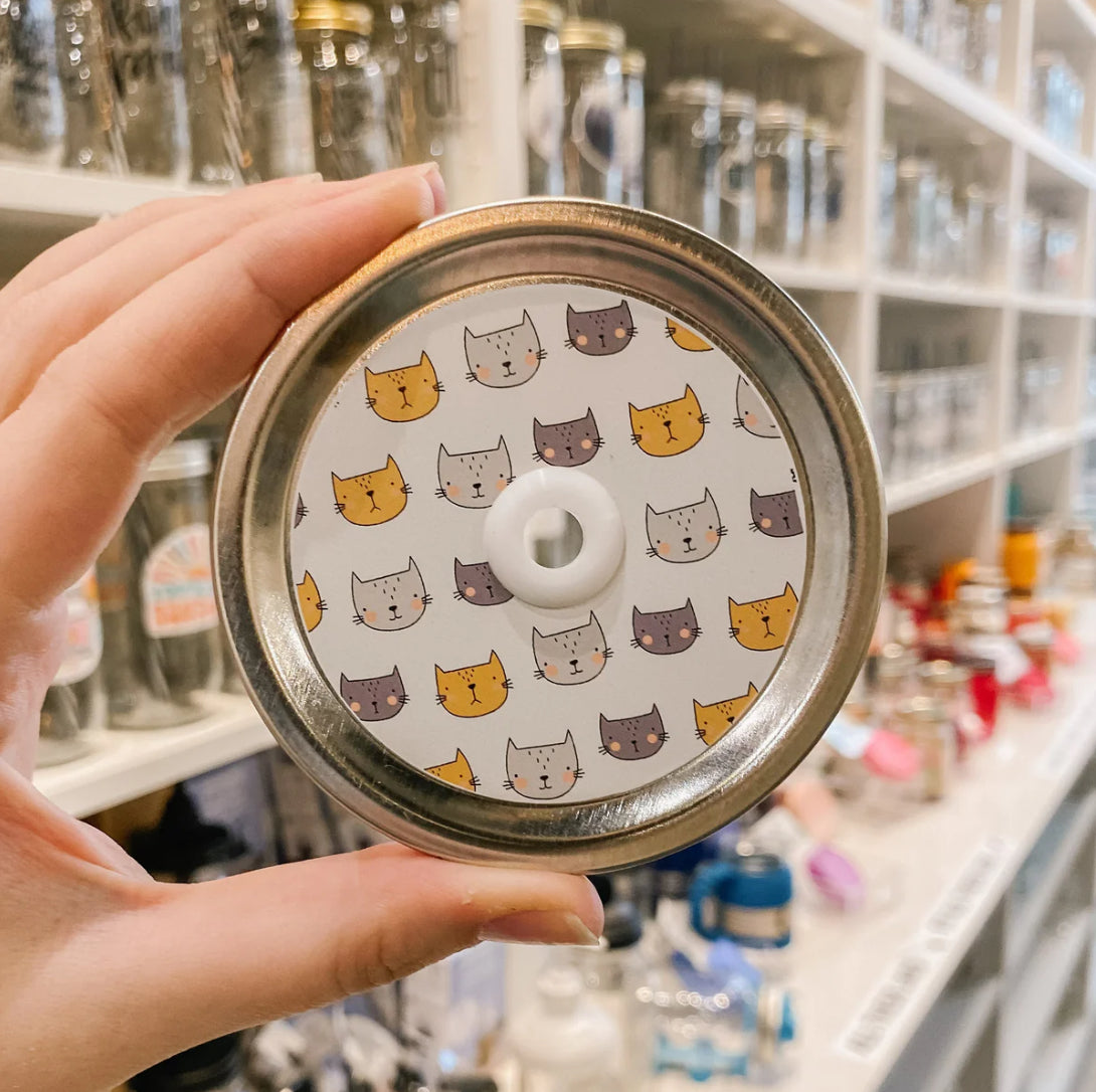 Hand holding mason jar patterned straw lid that has orange, light and dark grey cat heads on white 