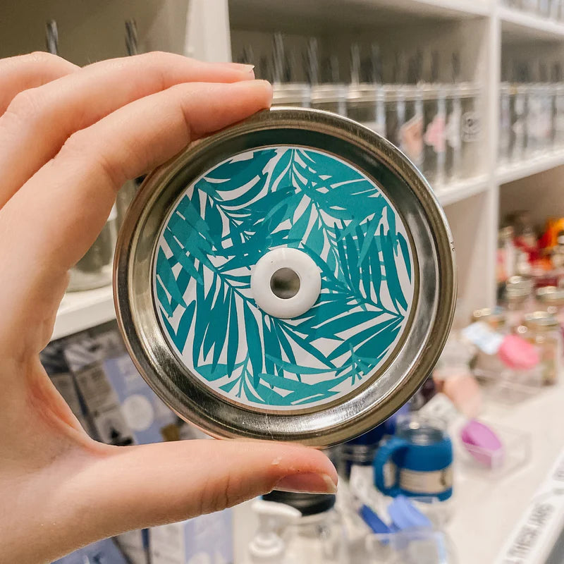 Hand holding mason jar patterned straw lid that has tropical teal leaves on white 