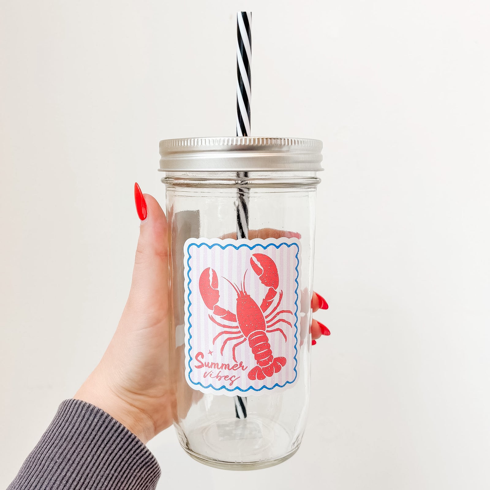 Hand holding mason jar tumbler that reads summer vibes in red ink with lobster design on pale pink and white striped background with blue border 
