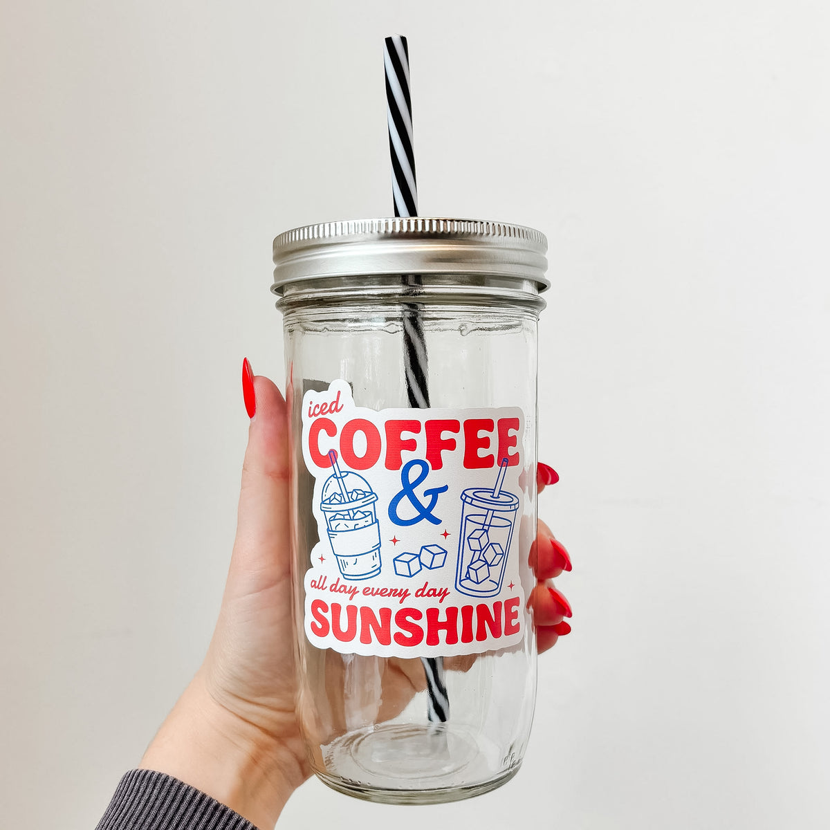 Hand holding mason jar tumbler that reads iced coffee & sunshine all day every day in red ink with blue iced drink designs on white background 