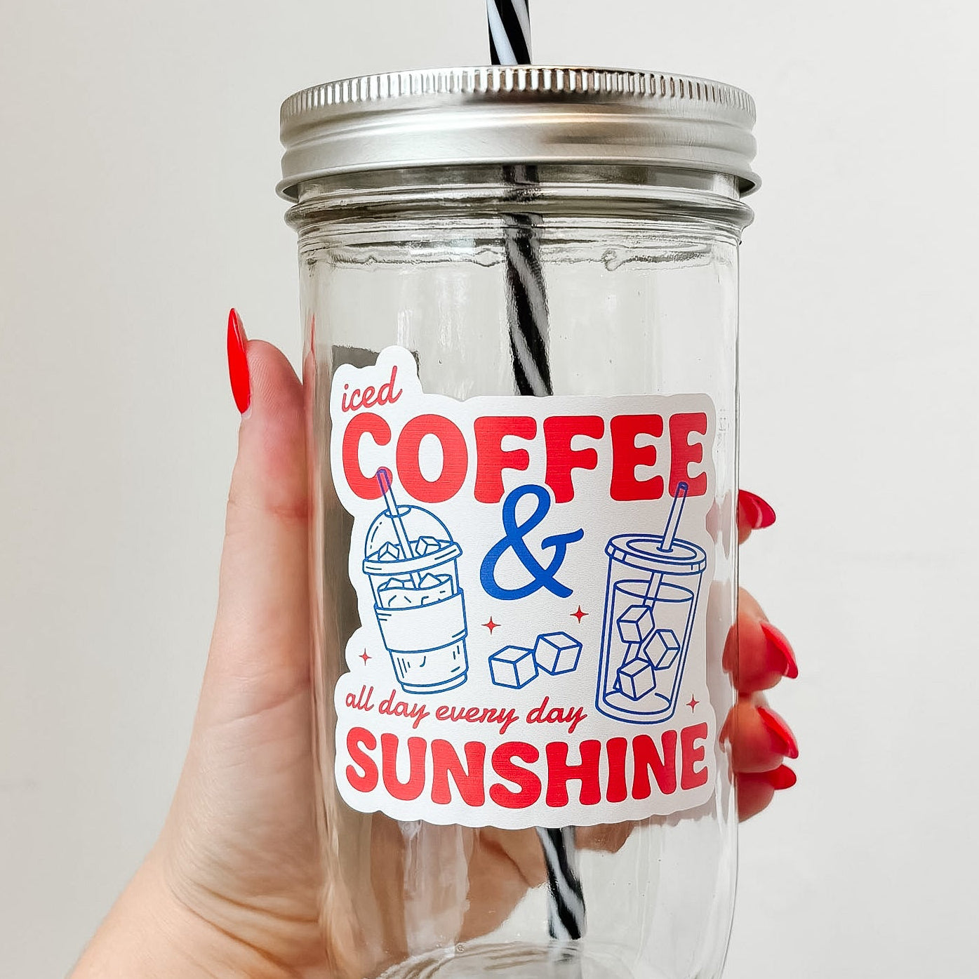 Hand holding mason jar tumbler that reads iced coffee & sunshine all day every day in red ink with blue iced drink designs on white background 