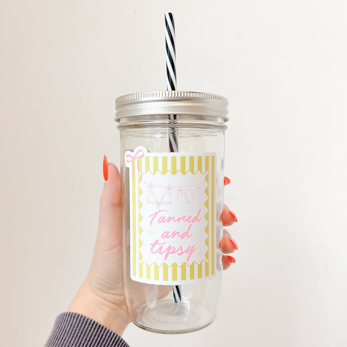 Hand holding mason jar tumbler that reads tanned and tipsy in pink font with yellow striped background and pink bow design 