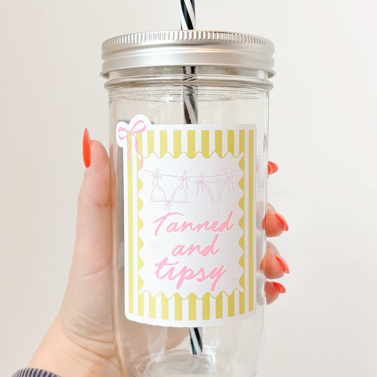 Hand holding mason jar tumbler up close that reads tanned and tipsy in pink font with yellow striped background and pink bow design 