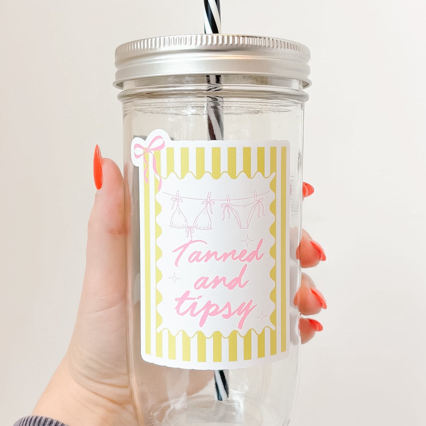 Hand holding mason jar tumbler that reads tanned and tipsy in pink font with yellow striped background and pink bow design 