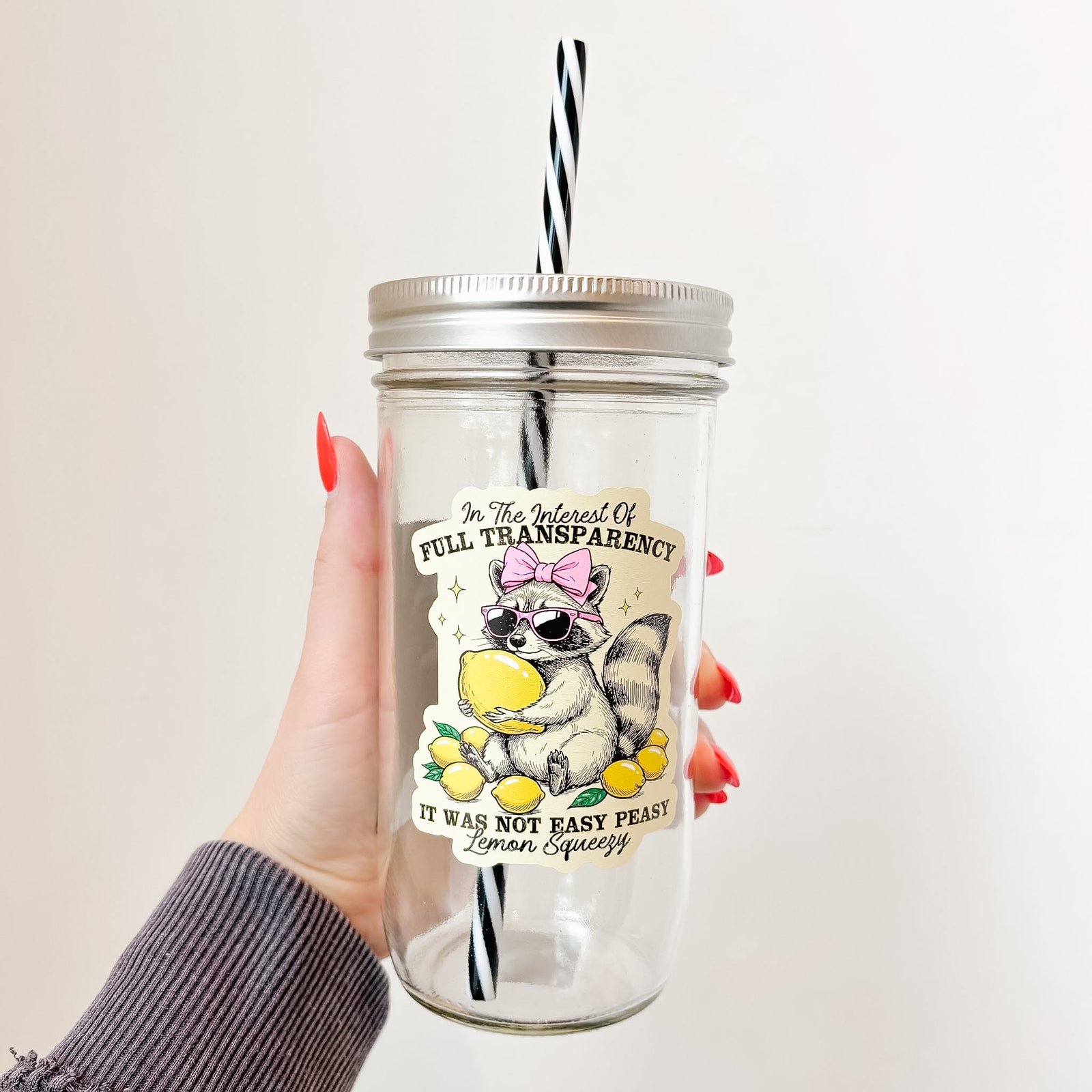 Hand holding mason jar tumbler that reads in the interest of full transparency it was not easy peasy lemon squeezy with a raccoon wearing pink sunglasses and bow holding a lemon and surrounded by lemons design on pale yellow background 