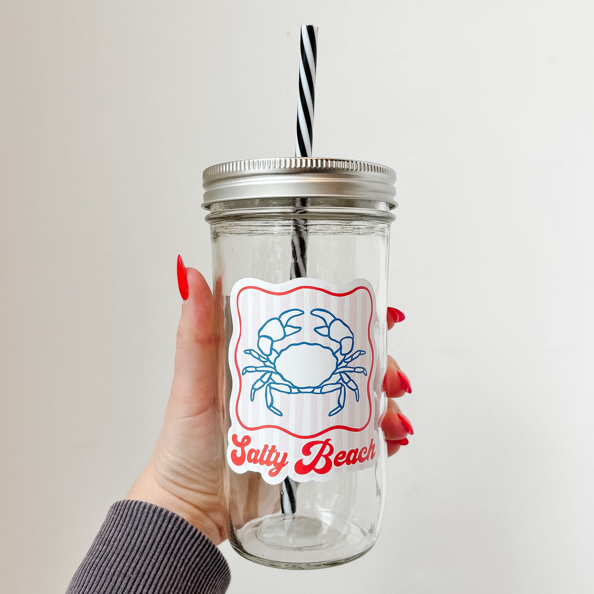 Hand holding mason jar tumbler that reads salty beach in red ink with blue crab design and red striped background with red border