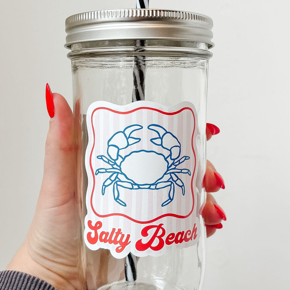 Hand holding mason jar tumbler up close that reads salty beach in red ink with blue crab design and red striped background with red border