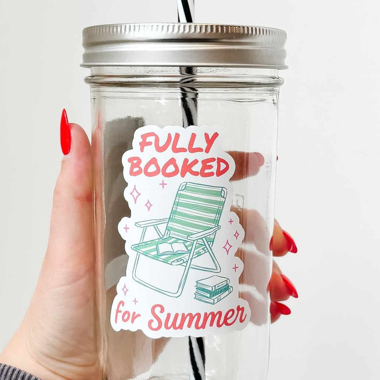 Hand holding mason jar tumbler that reads fully booked for summer in red font with green beach hair and stack of books