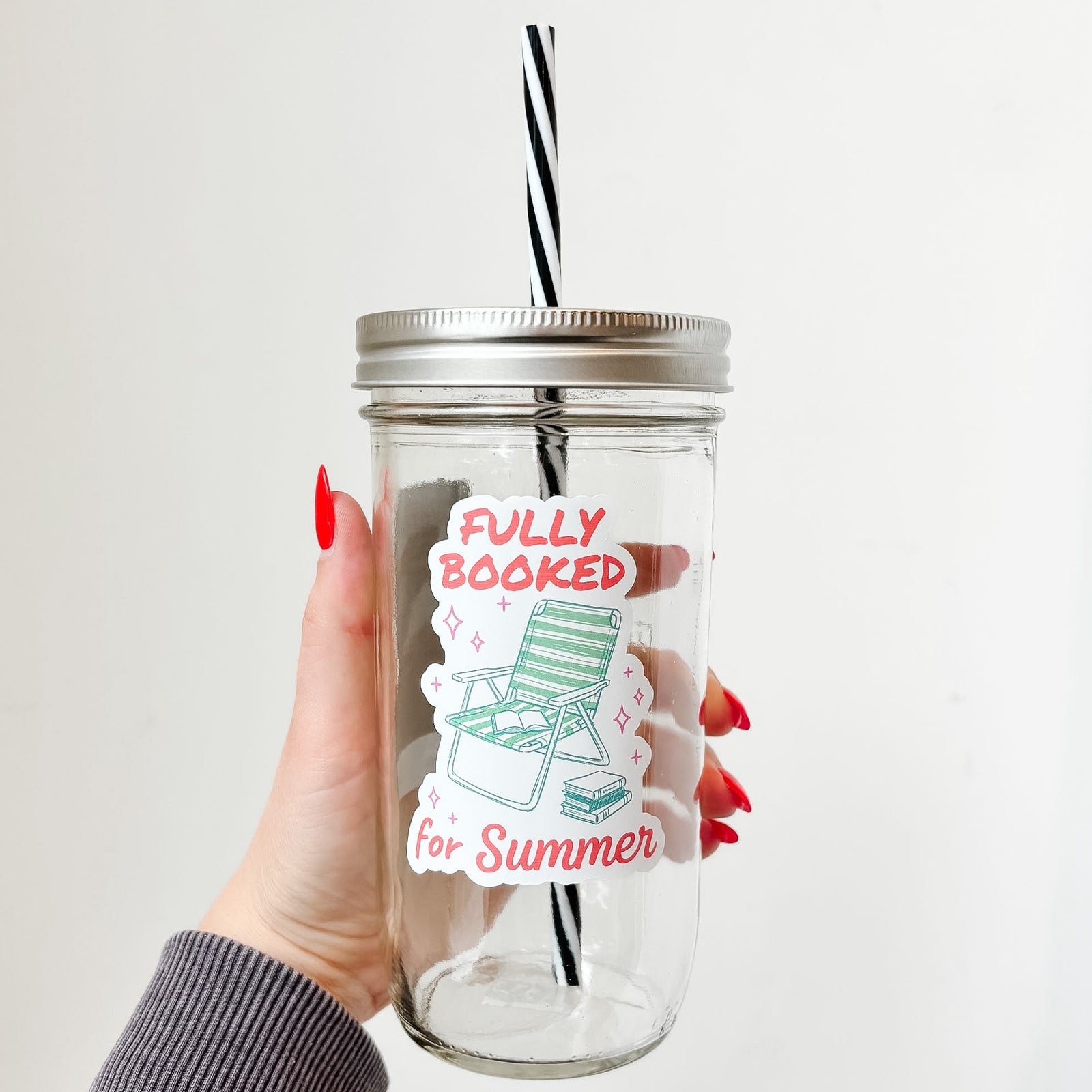 Hand holding mason jar tumbler that reads fully booked for summer in red font with green beach hair and stack of books