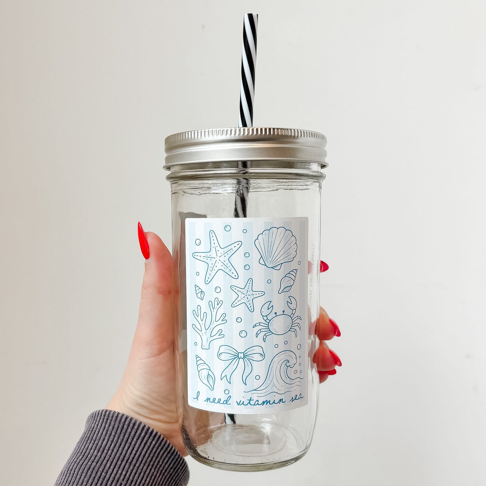 Hand holding mason jar tumbler that reads i need vitamin sea with starfish, shell, crab, coral and other ocean designs on pale blue and white striped background 