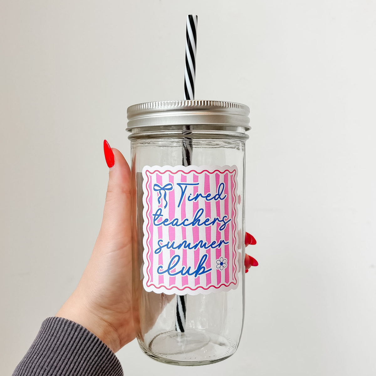 Hand holding mason jar tumbler that reads tired teachers club in blue ink with blue bow and flower on pink and white stripe background with red border 