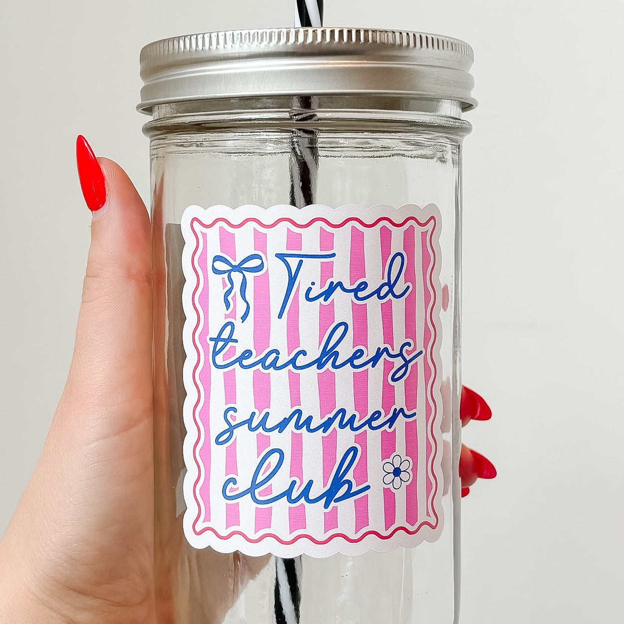 Hand holding mason jar tumbler that reads tired teachers club in blue ink with blue bow and flower on pink and white stripe background with red border 