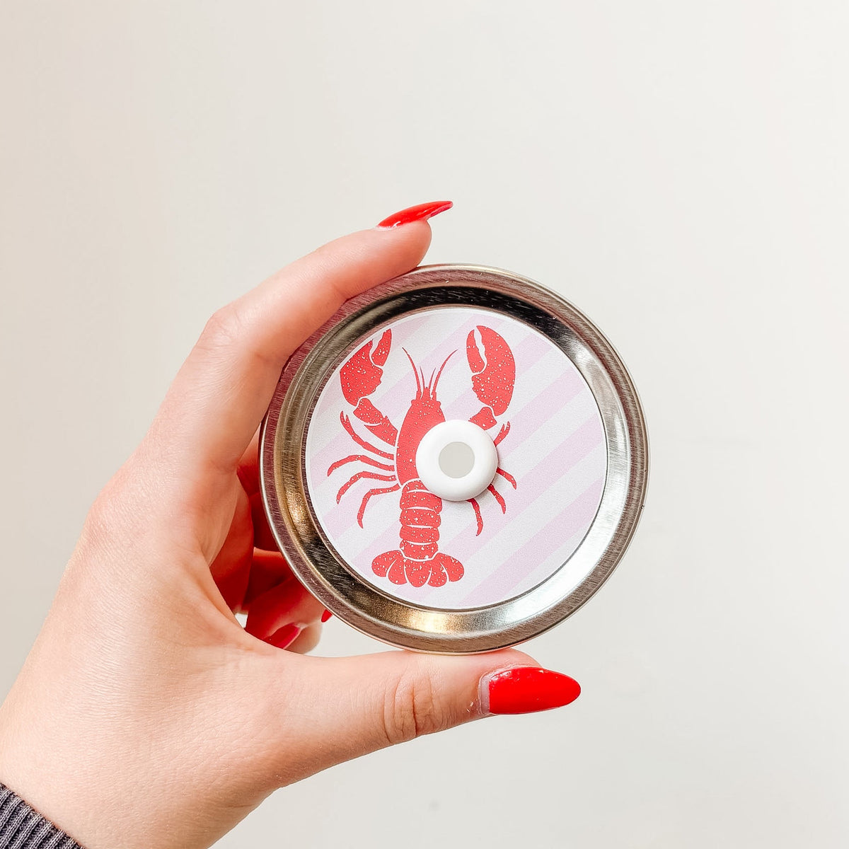 Hand holding mason jar patterned straw lid that has red lobster design on pale pink and white diagonal stripe background 