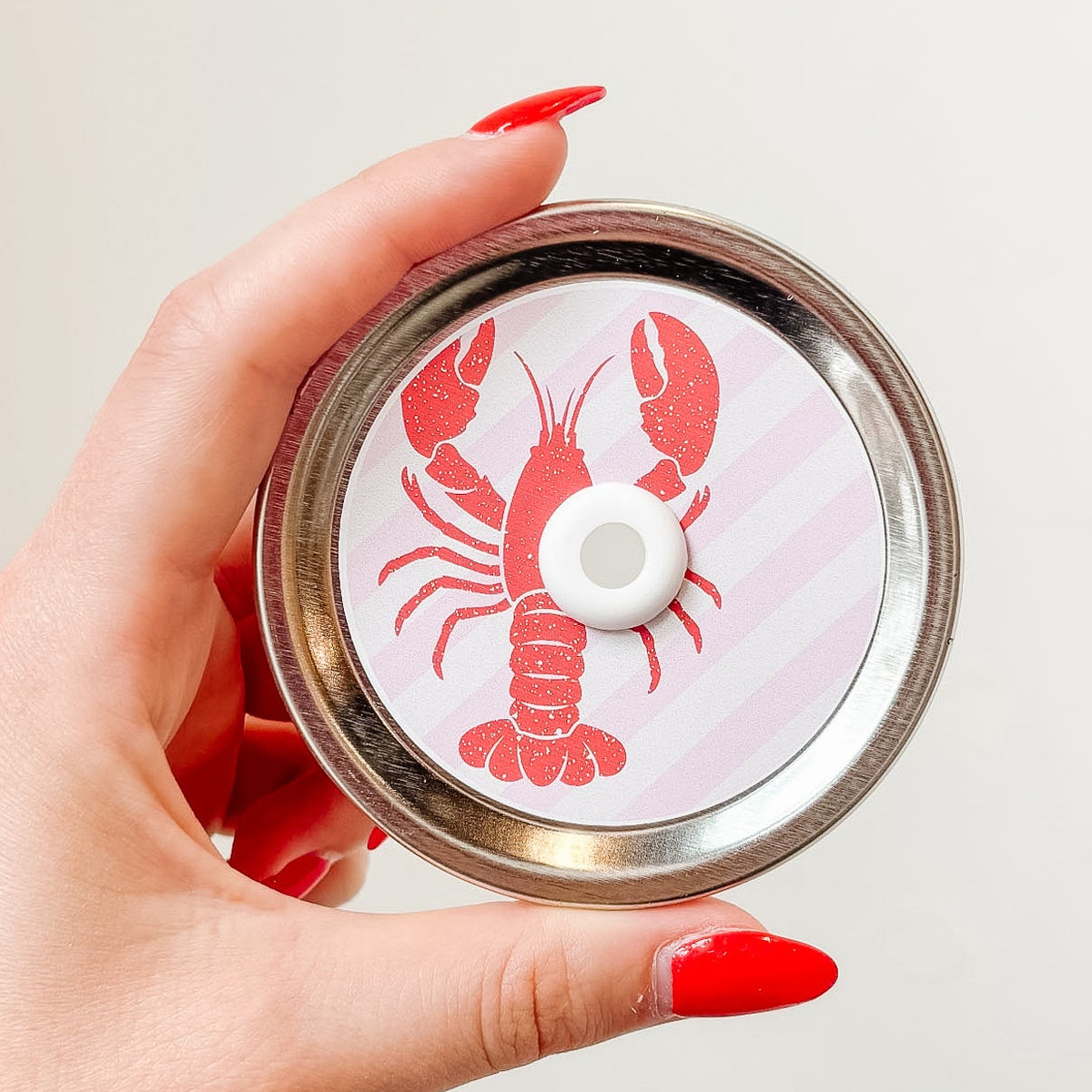 Hand holding mason jar patterned straw lid up close that has red lobster design on pale pink and white diagonal stripe background 