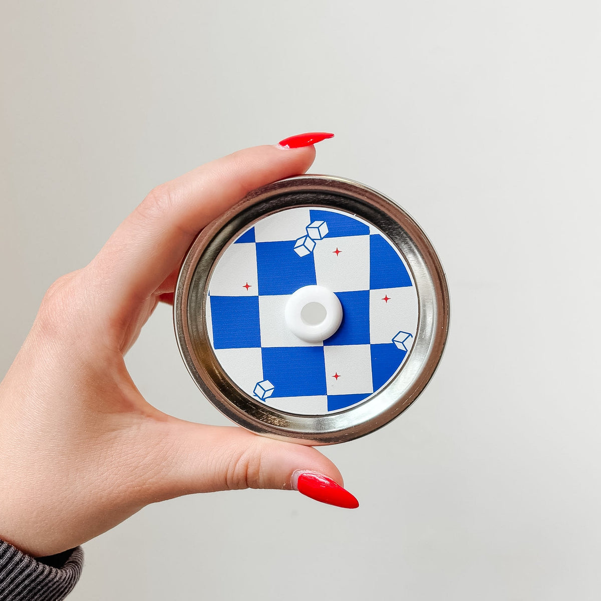 Hand holding a mason jar patterned straw lid that has blue and white checkered design with little iced cubes and tiny red stars