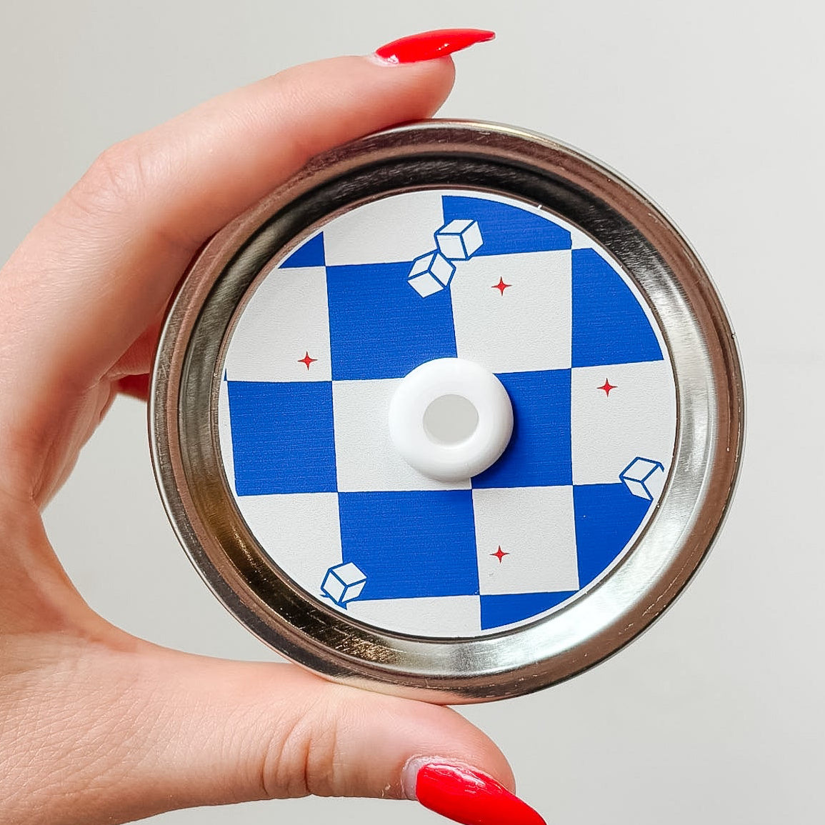 Hand holding a mason jar patterned straw lid up close that has blue and white checkered design with little iced cubes and tiny red stars