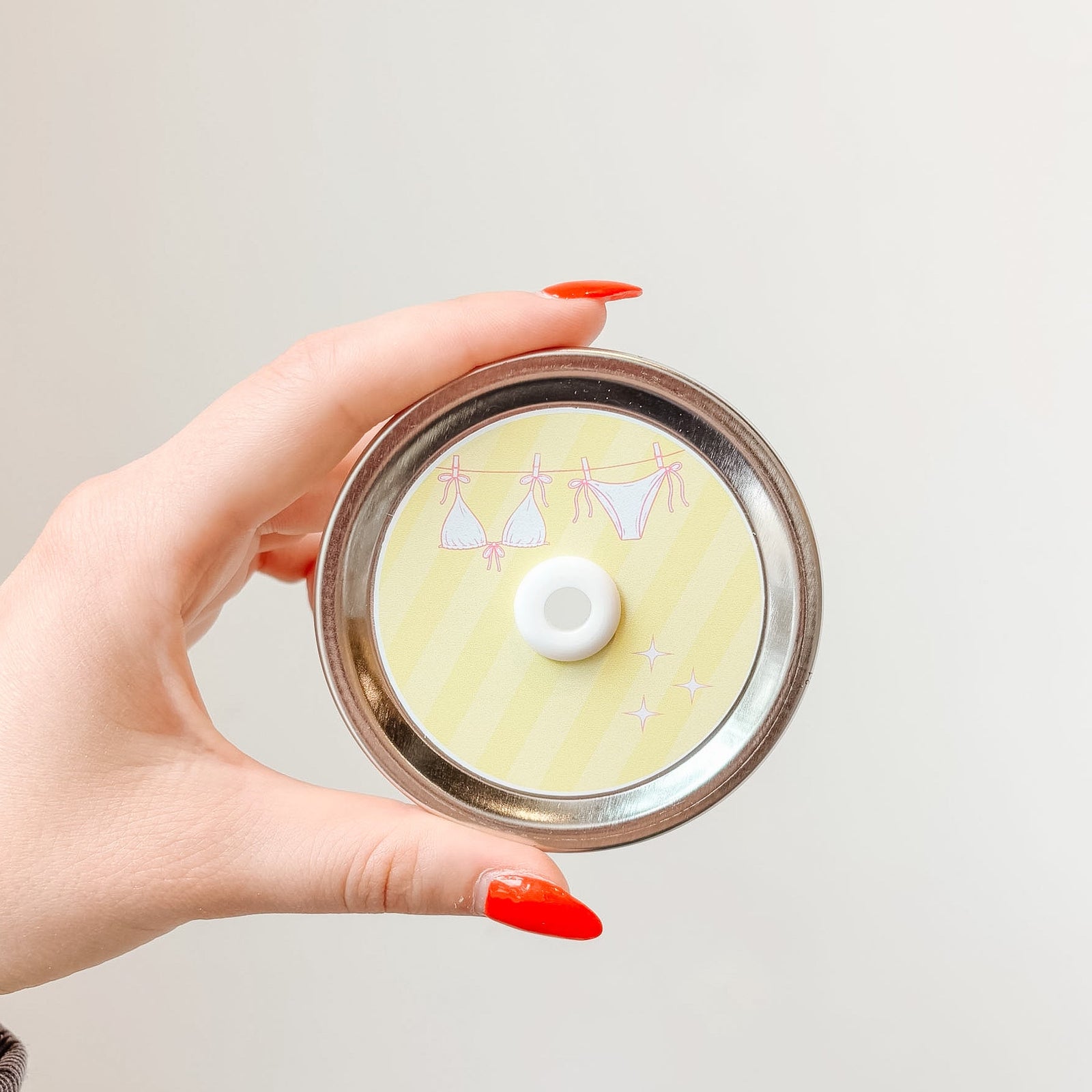 Hand holding mason jar patterned straw lid that has a pink bikini on a clothesline design on light yellow striped background 