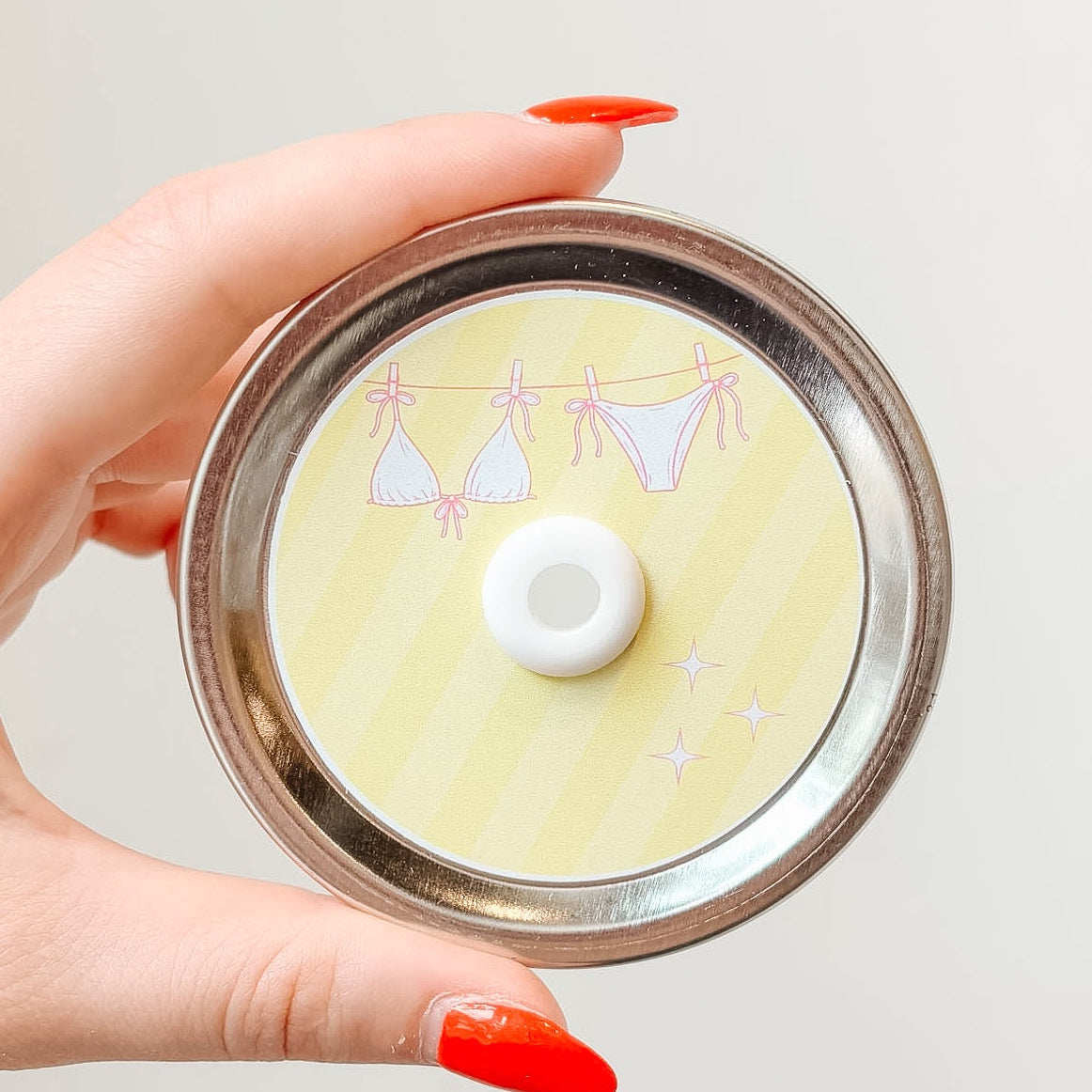 Hand holding mason jar patterned straw lid up close that has a pink bikini on a clothesline design on light yellow striped background