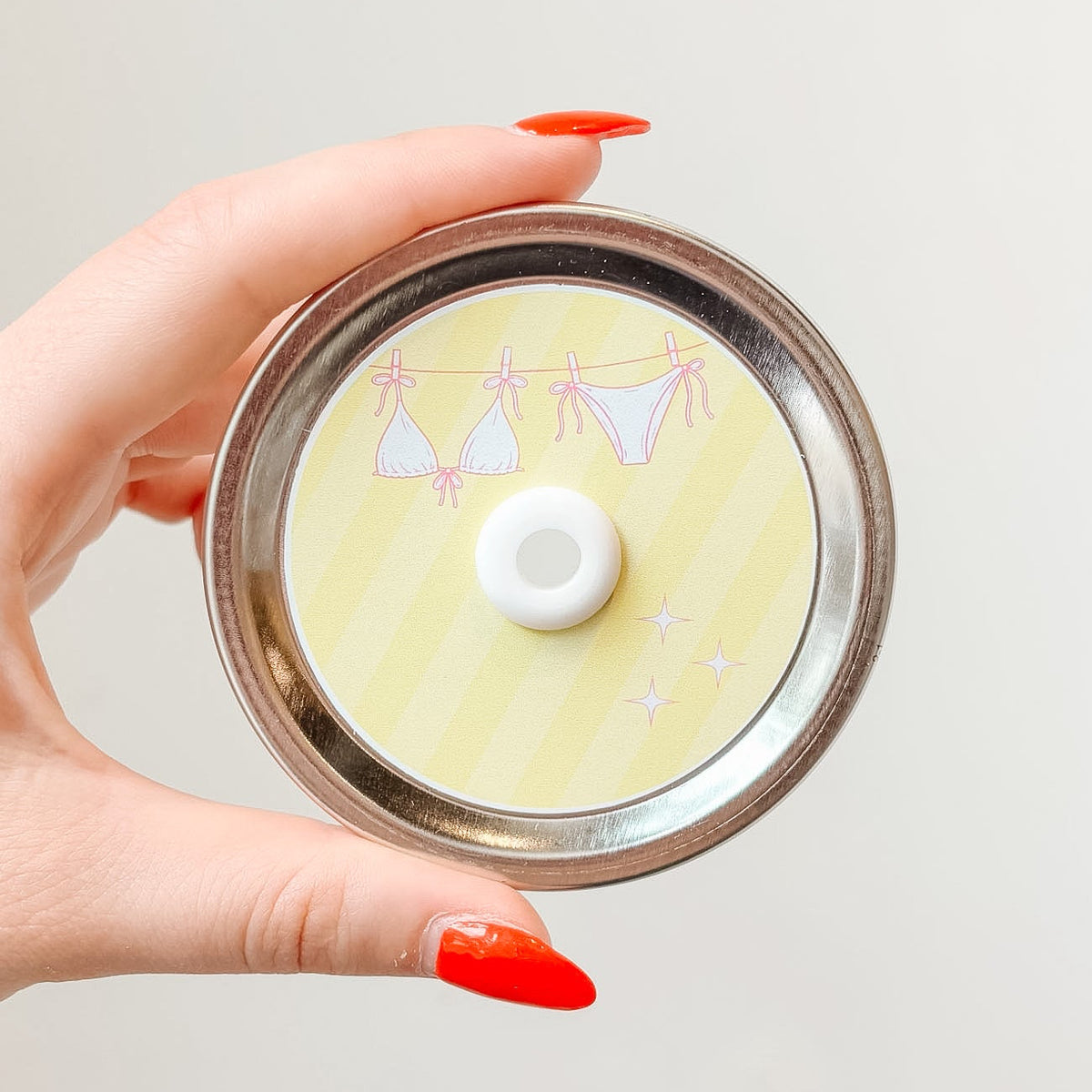 Hand holding mason jar patterned straw lid up close that has a pink bikini on a clothesline design on light yellow striped background