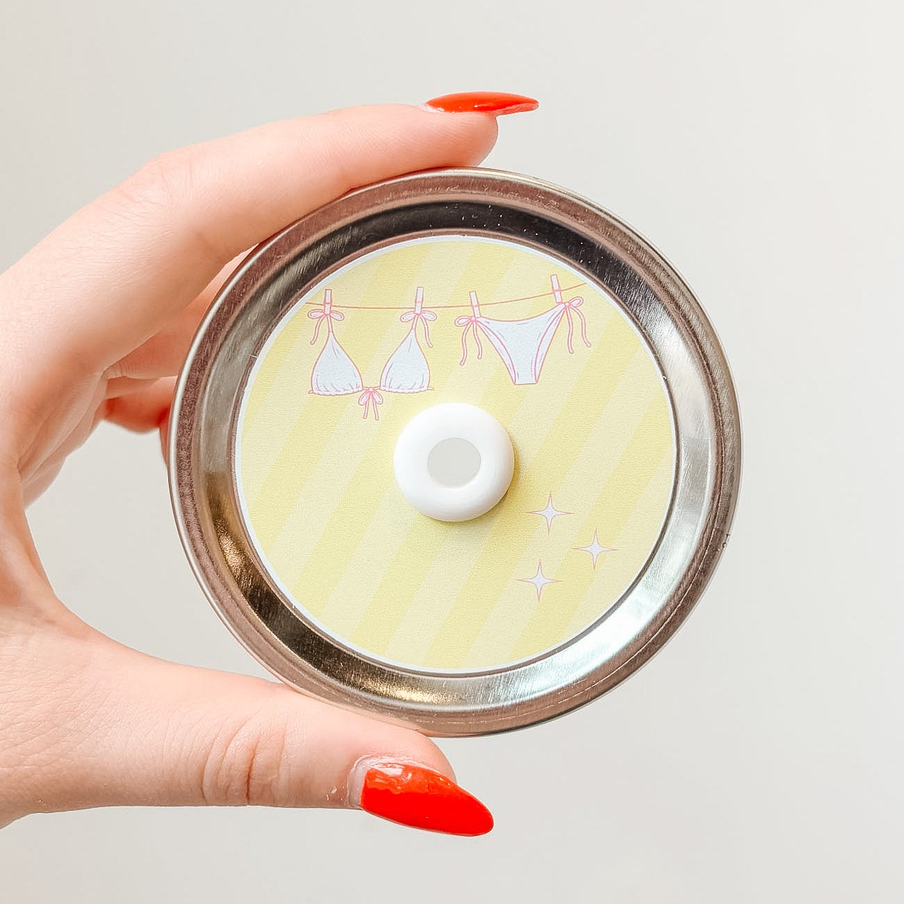 Hand holding mason jar patterned straw lid up close that has a pink bikini on a clothesline design on light yellow striped background