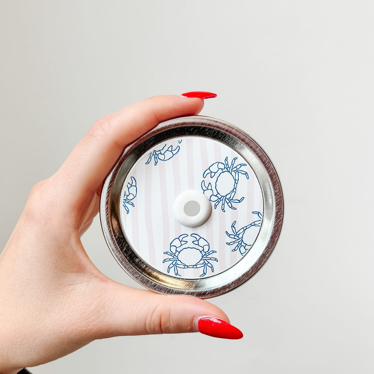Hand holding mason jar patterned straw lid that has blue crab designs on pale pink and white striped background