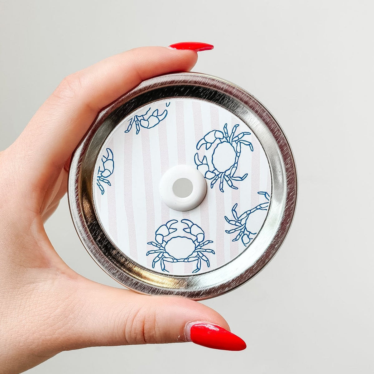 Hand holding mason jar patterned straw lid up close that has blue crab designs on pale pink and white striped background