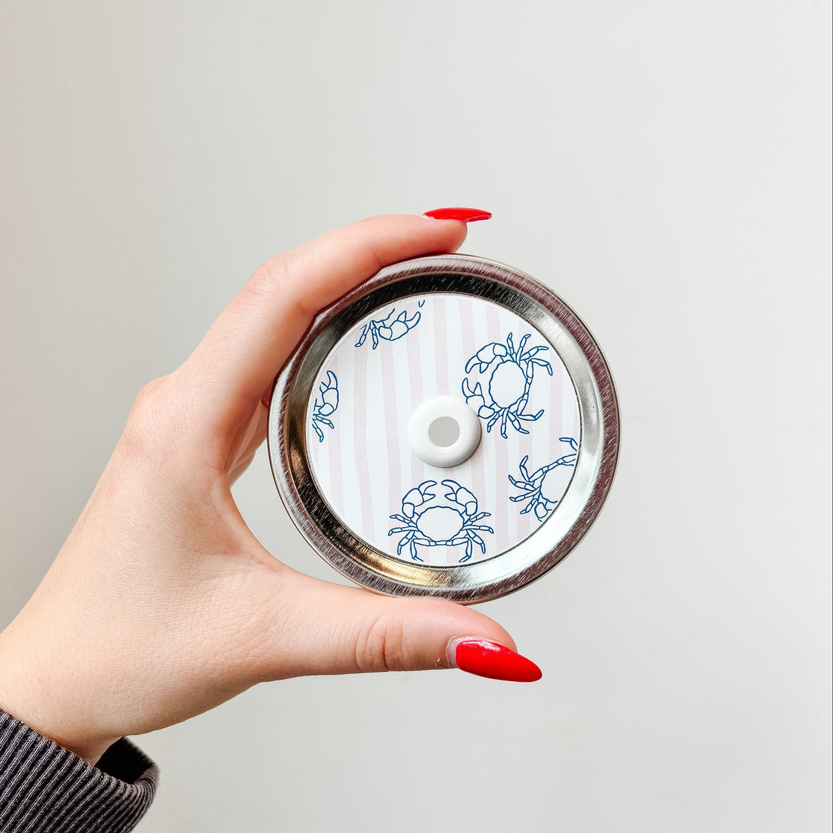 Hand holding mason jar patterned straw lid that has blue crab designs on pale pink and white striped background 