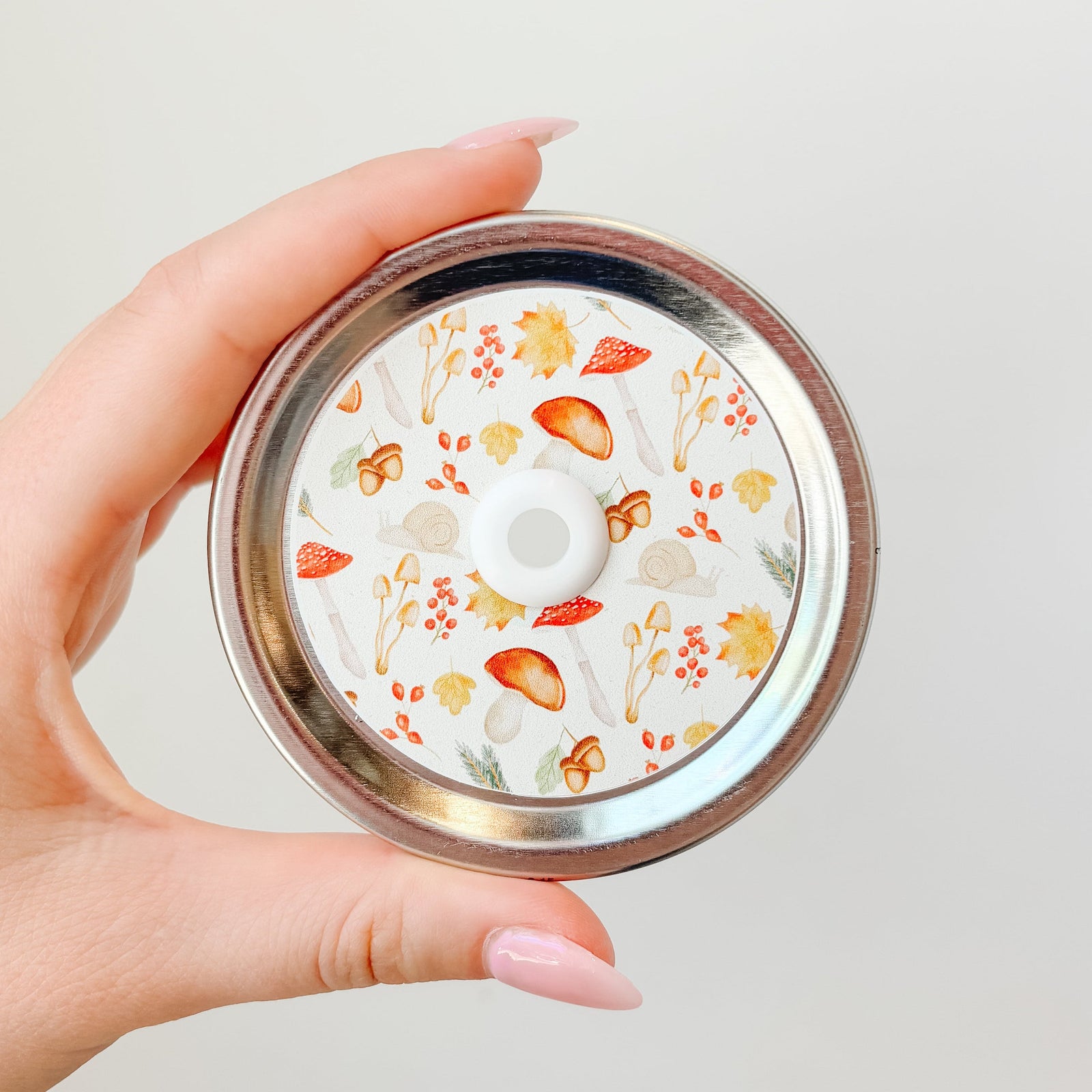 Hand holding mason jar patterned straw lid that has multi-autumn designs such as mushrooms, leaves and acorns on white background 