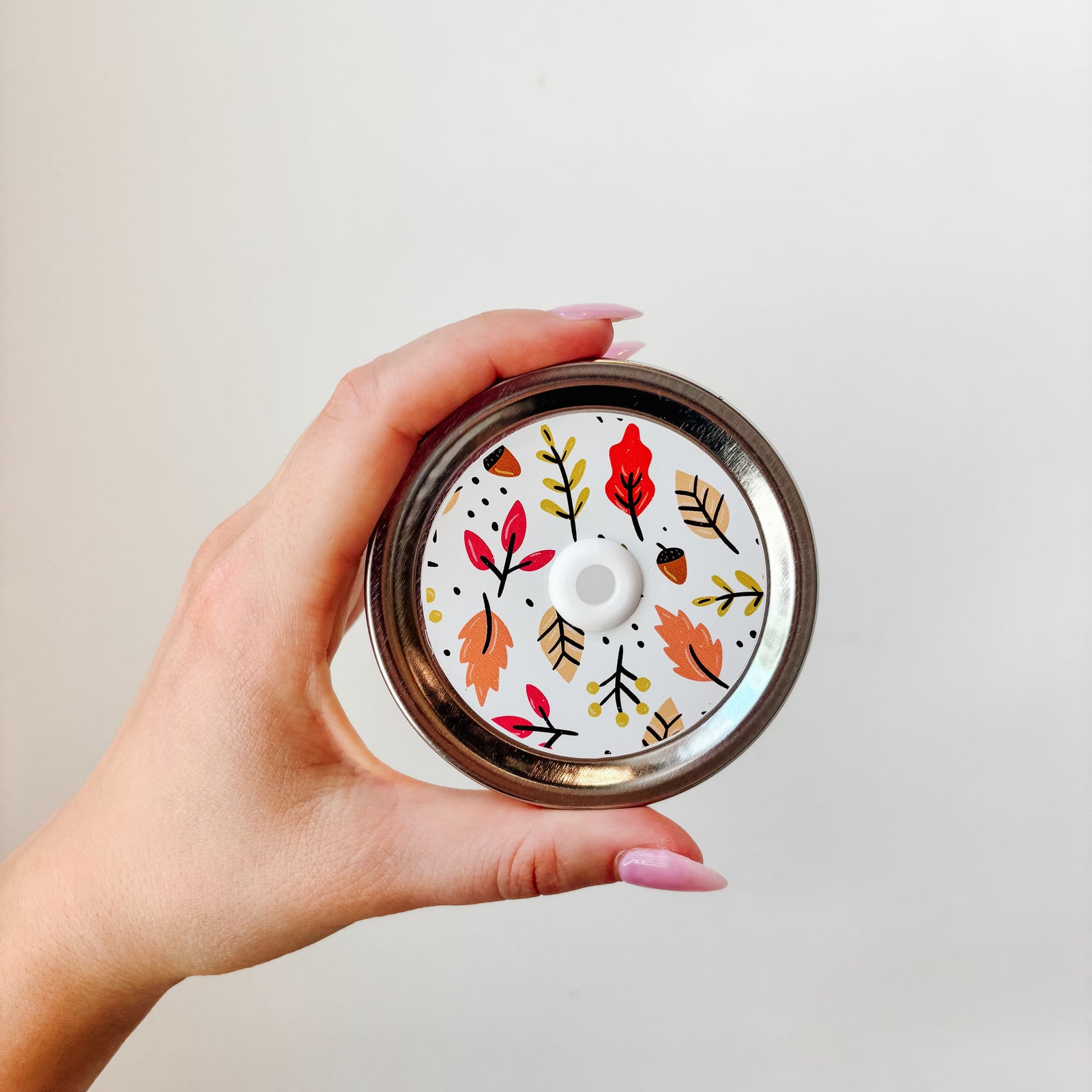 Hand holding mason jar patterned straw lid that had chunky multi-coloured leaves on white