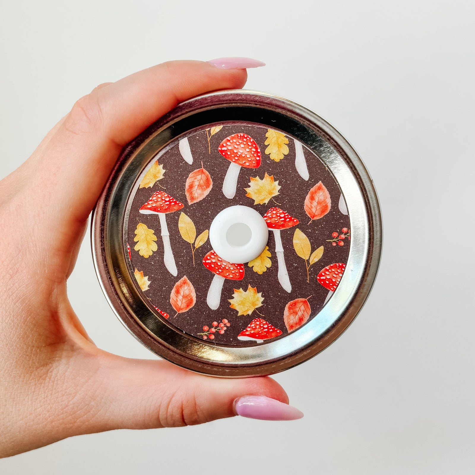 Hand holding mason jar patterned straw lid that has red mushrooms and multi-coloured leaves on dark brown background.