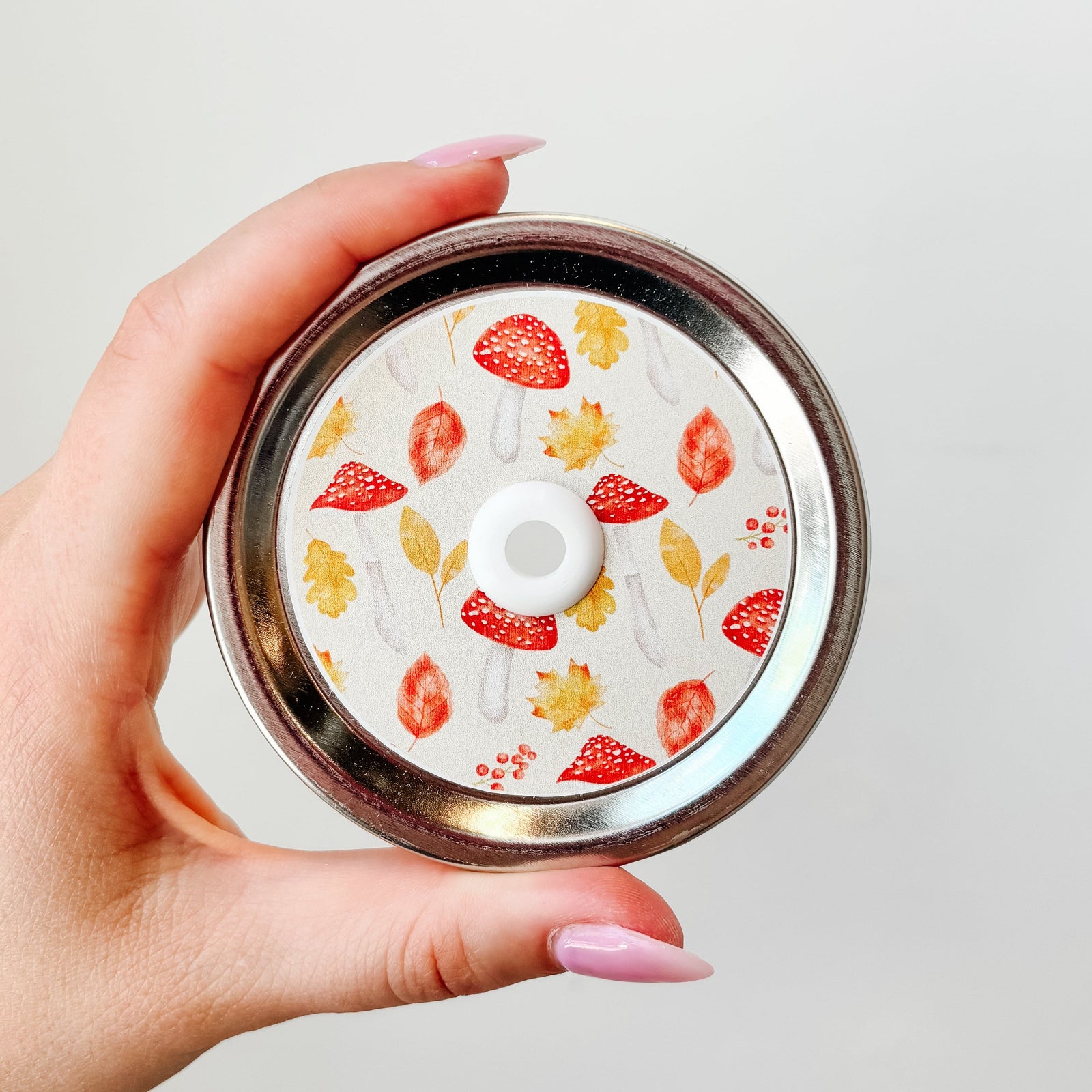 Hand holding mason jar patterned straw lid that has multi-autumn designs such as mushrooms and orange leaves on white background