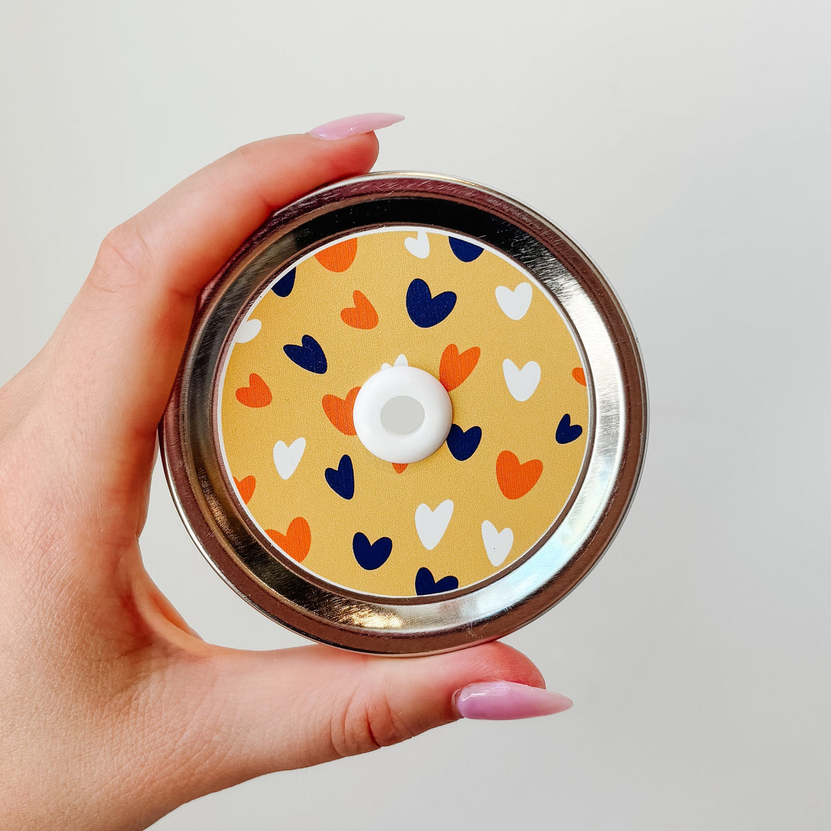 Hand holding mason jar patterned straw lid that has navy, white and orange hearts on light orange background. 