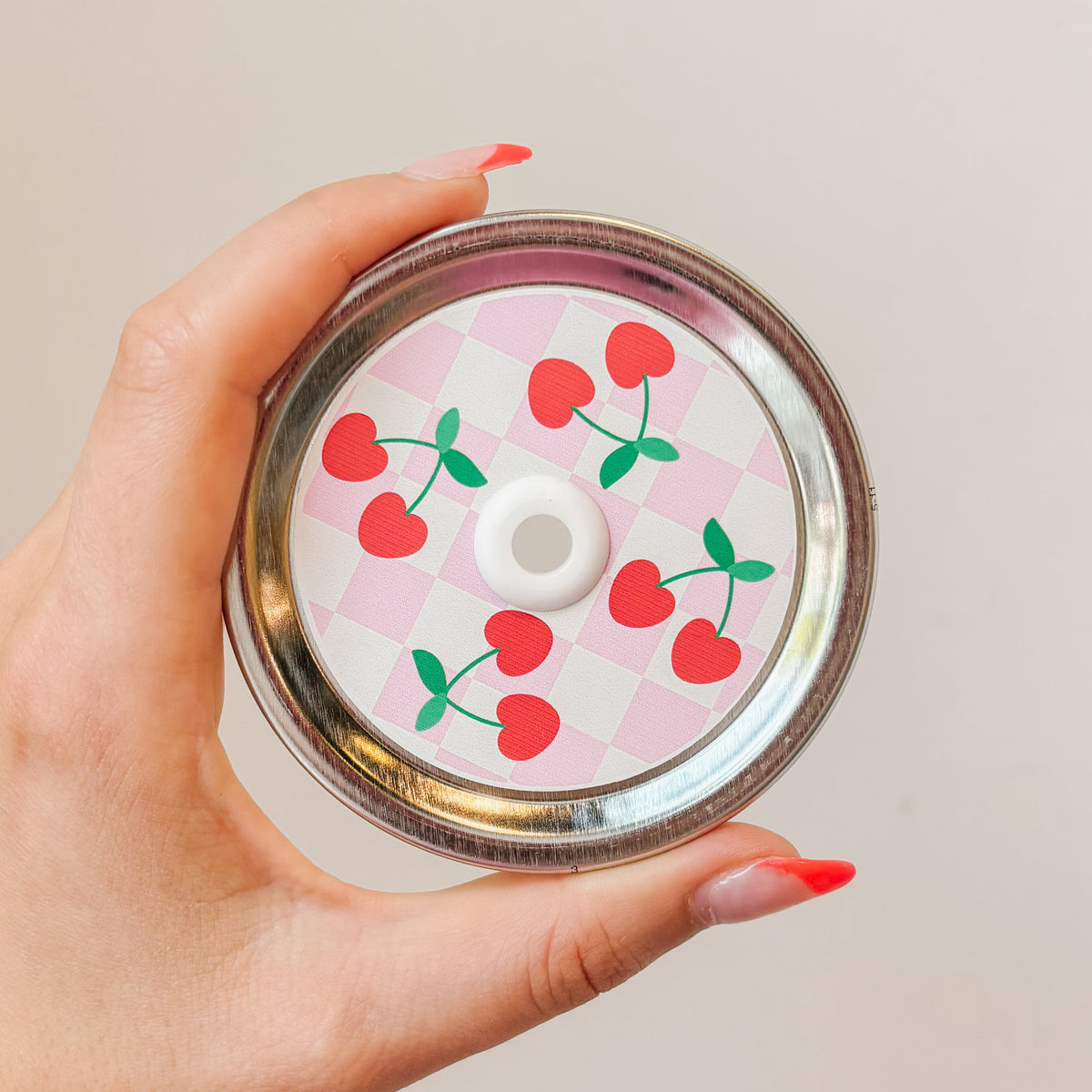 Hand holding a mason jar patterned straw lid with cherries on a white and light, pink checkered background
