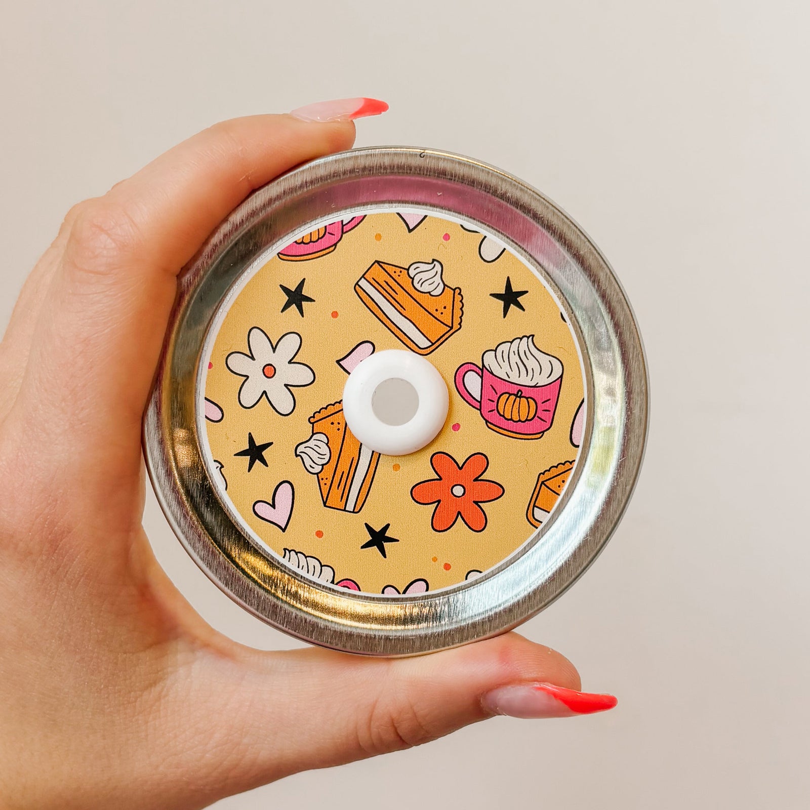 Hand holding a mason jar patterned straw lid that has pumpkin pie, mugs, florals and designs on light orange background 