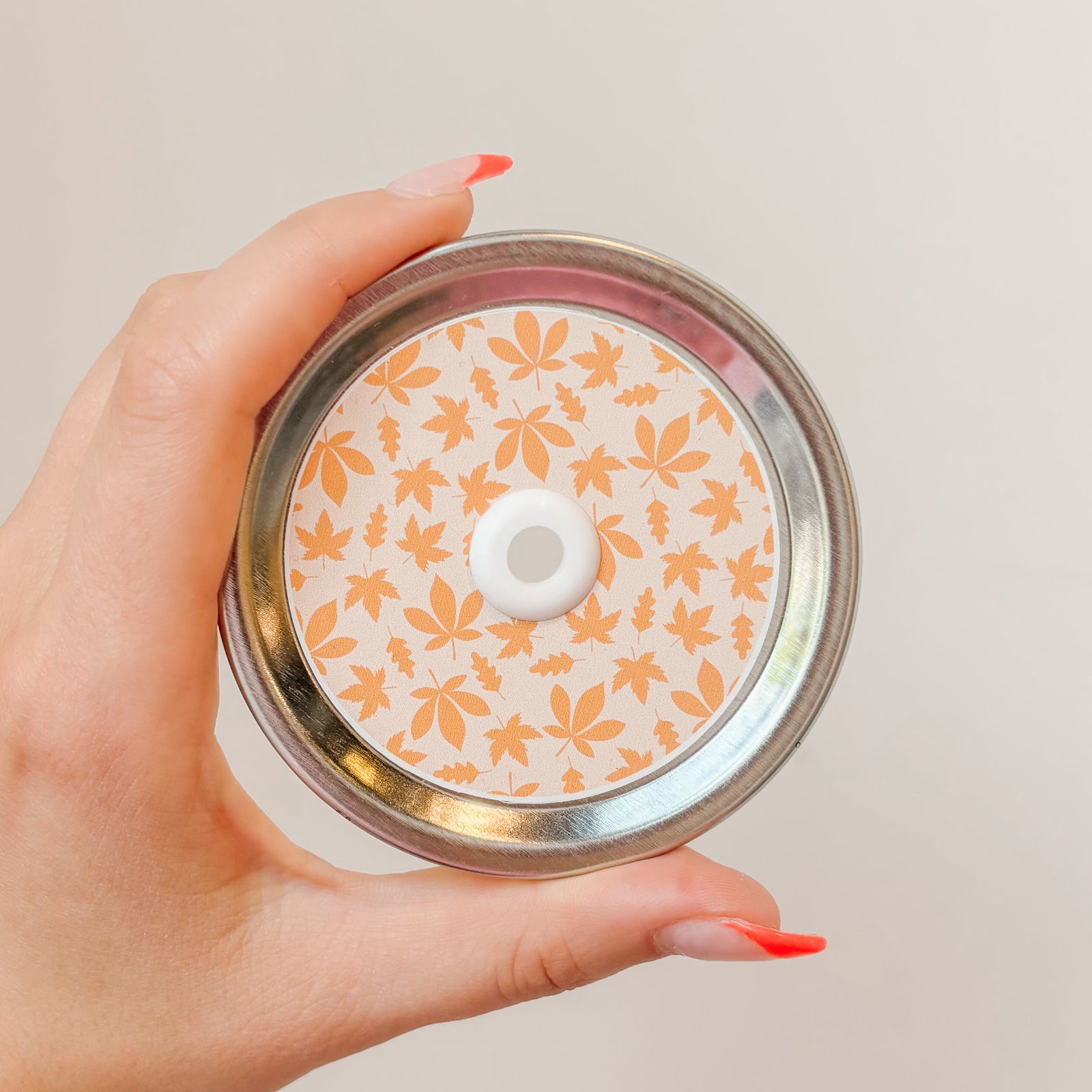 Hands holding mason jar patterned straw lid that has assorted orange leaves on light pale orange background