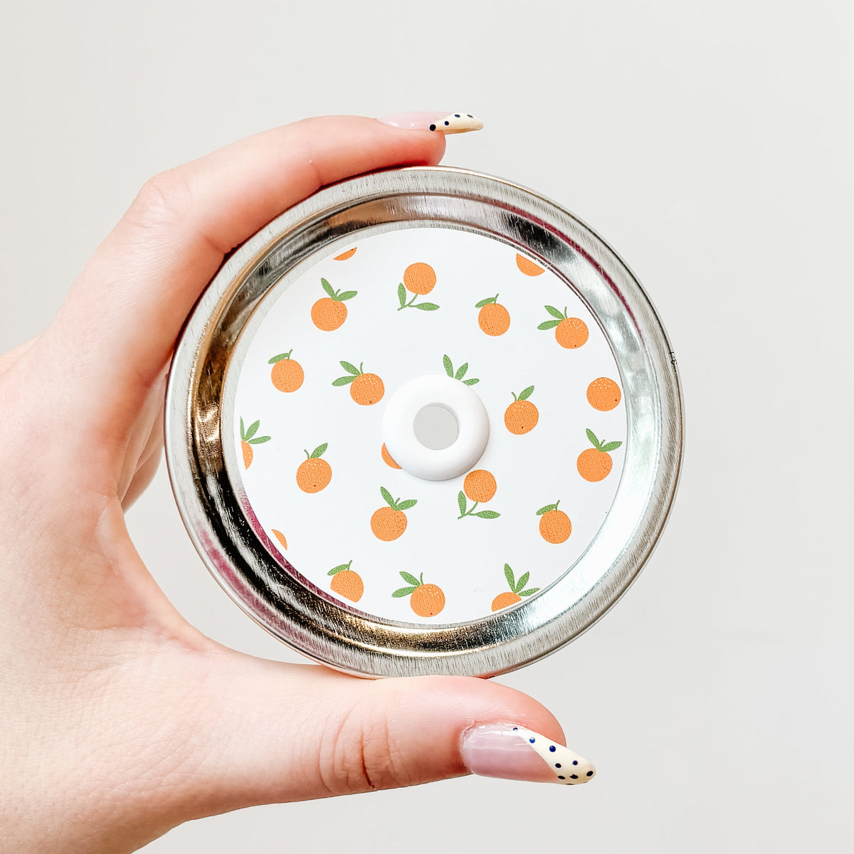 Hand holding a mason jar patterned straw lid with little oranges on a white background.