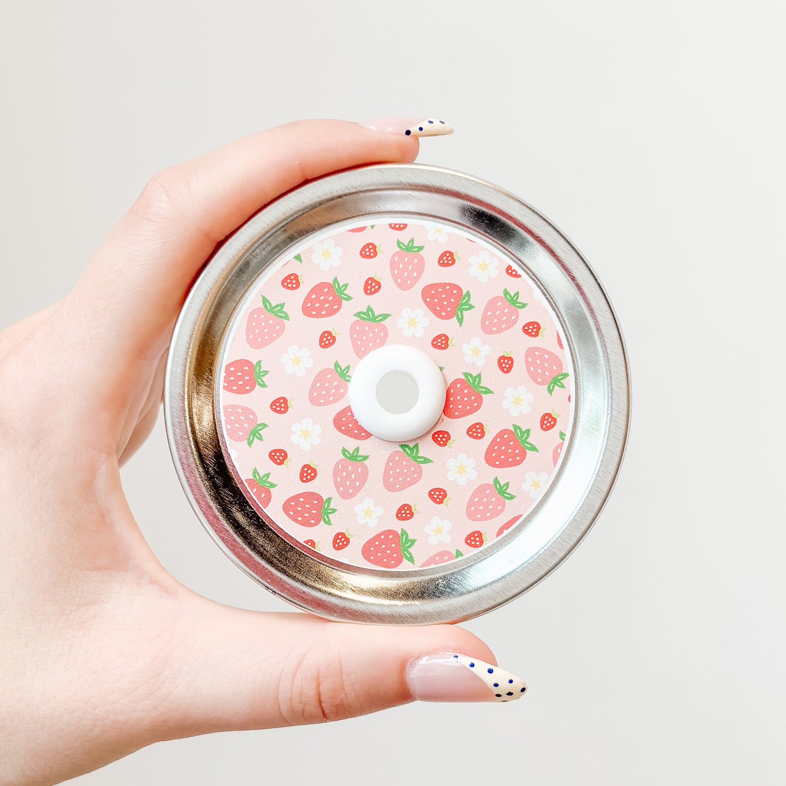 A hand holding a mason jar patterned straw lid with assorted strawberries and flowers on a light pink background
