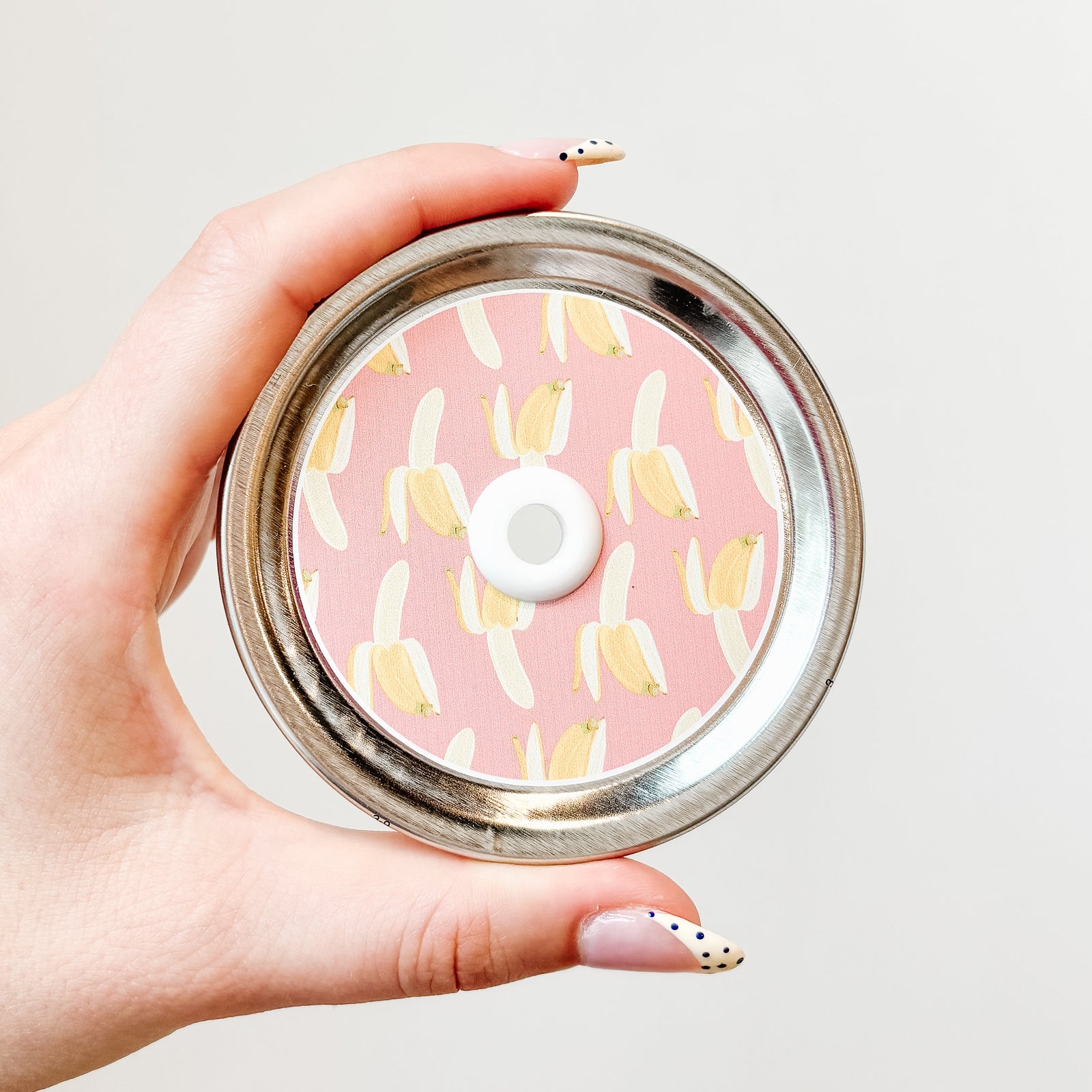 A hand holding a mason jar patterned straw lid with bananas on a pink background