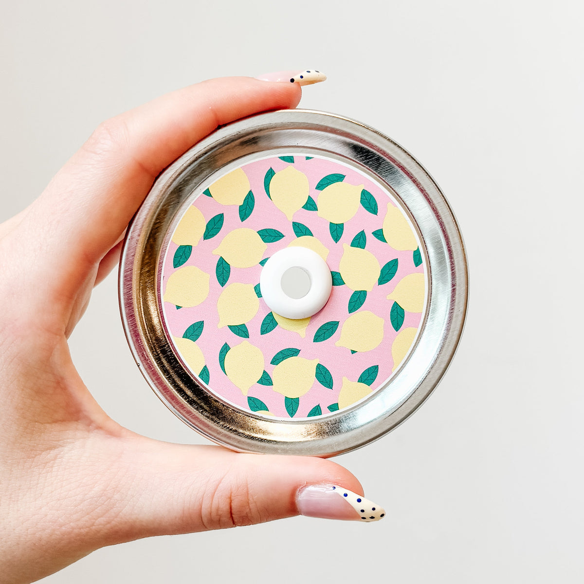A hand holding a mason jar patterned straw lid with lemons on a light pink background