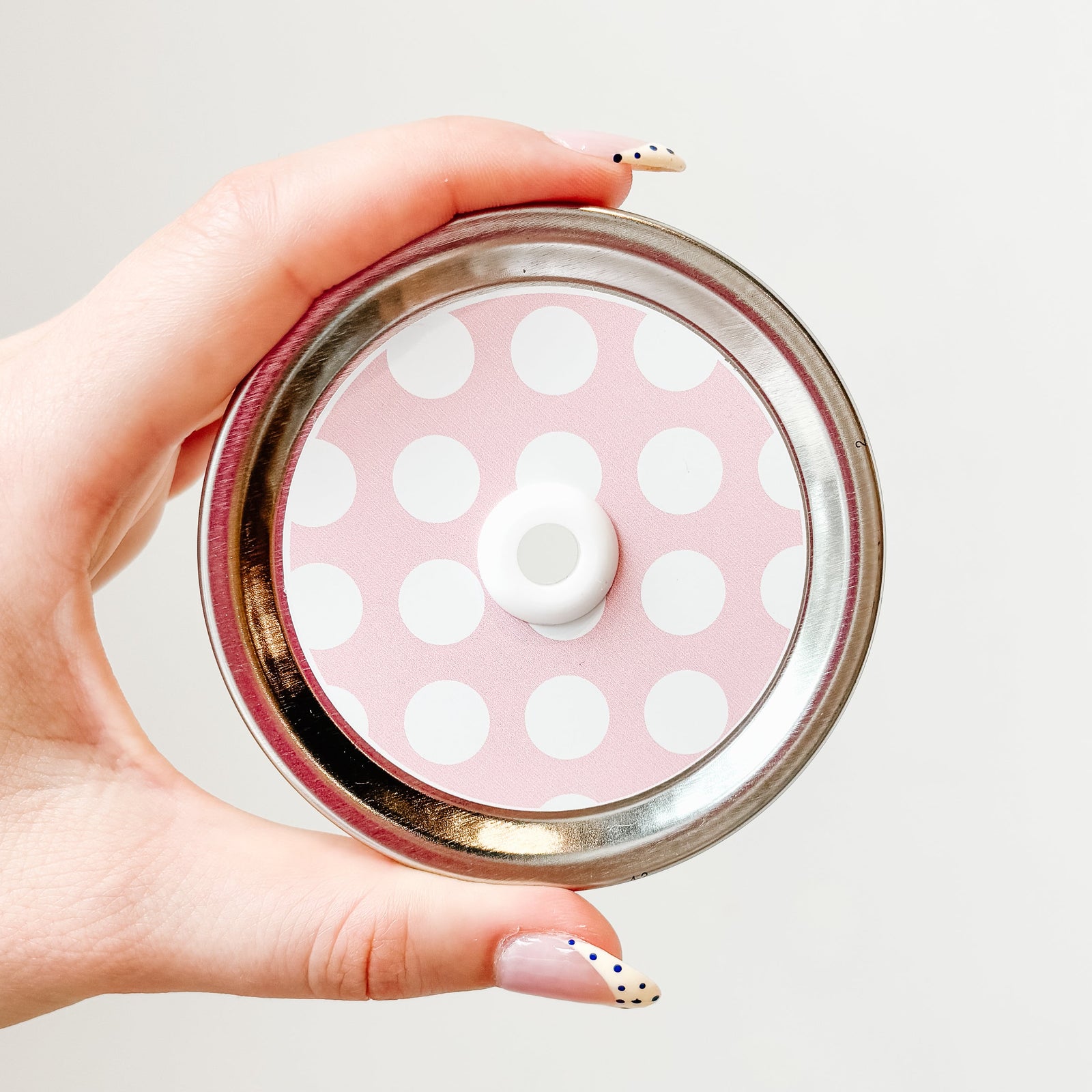 Hand holding mason jar patterned straw lid that has large white dots on light pink background