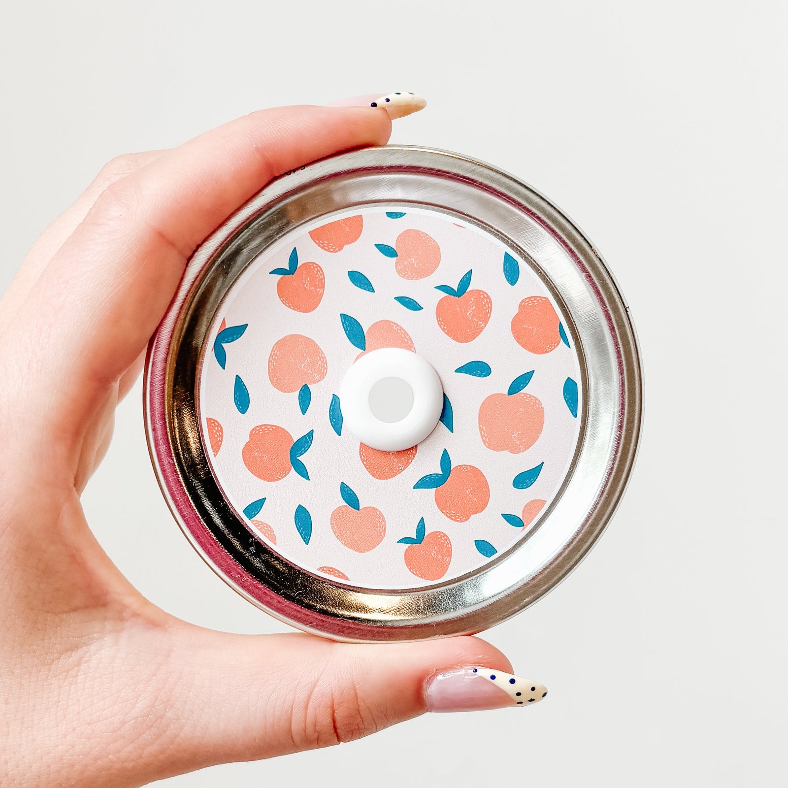 Hand holding a patterned mason jar straw lid that has peaches with teal leaves on an off-white background
