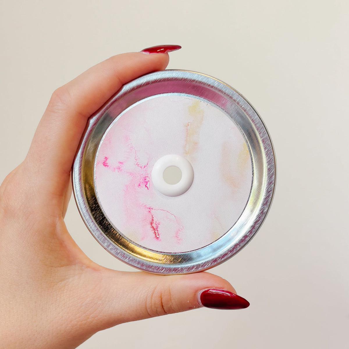 A hand holding a mason jar straw lid that has a watercolour pink and yellow design.