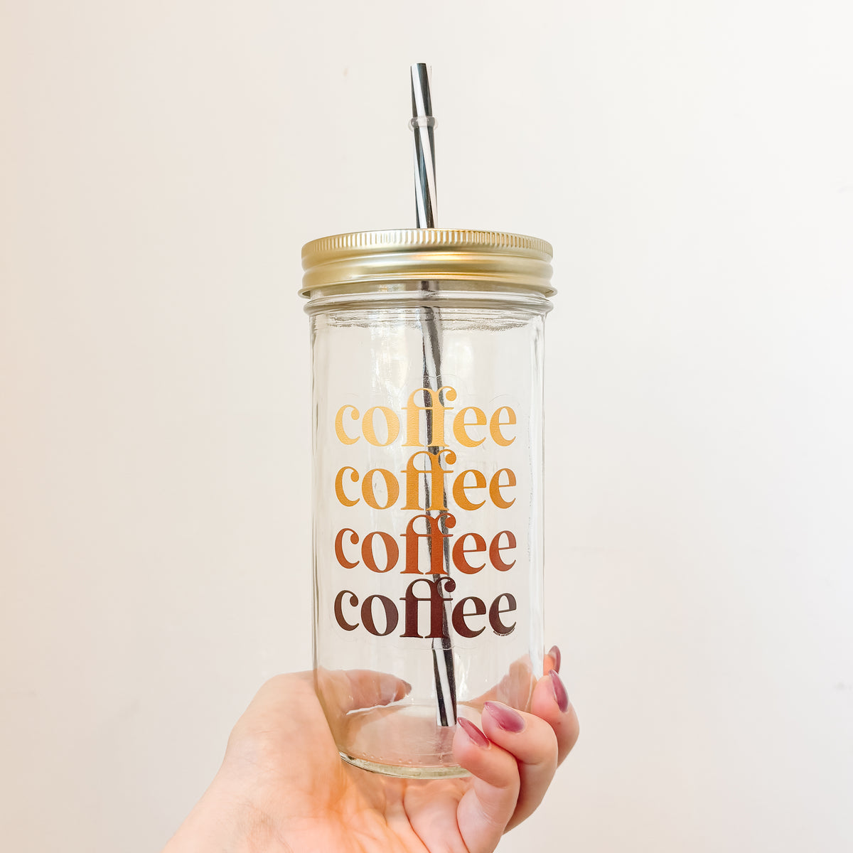 A hand holding a clear glass mason jar tumbler with a gold metal lid and a black-and-white striped shaw. The glass features the word 'coffee' repeated vertically in a retro gradient from gold to orange to brown.