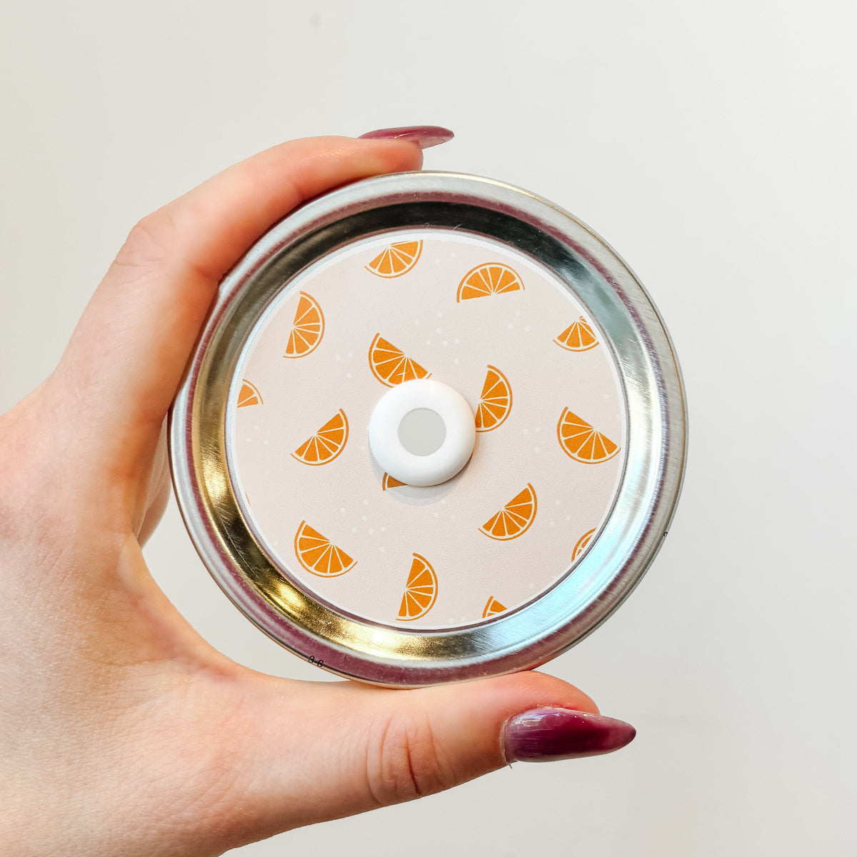 hand holding a mason jar straw lid that has small orange slices on light pale orange background.