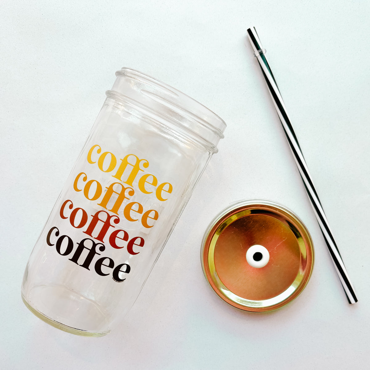 A clear glass tumbler with the word 'coffee' repeated vertically in yellow, orange, and brown, shown with a metallic gold straw lid and a black-and-white striped straw.