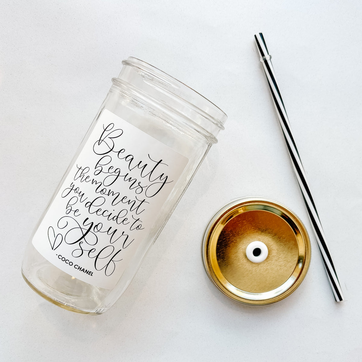 A clear glass tumbler with a white script label reading "Beauty begins the moment you decide to be yourself- Coco Chanel", shown on a white background with a gold straw lid and a black-and-white straw.