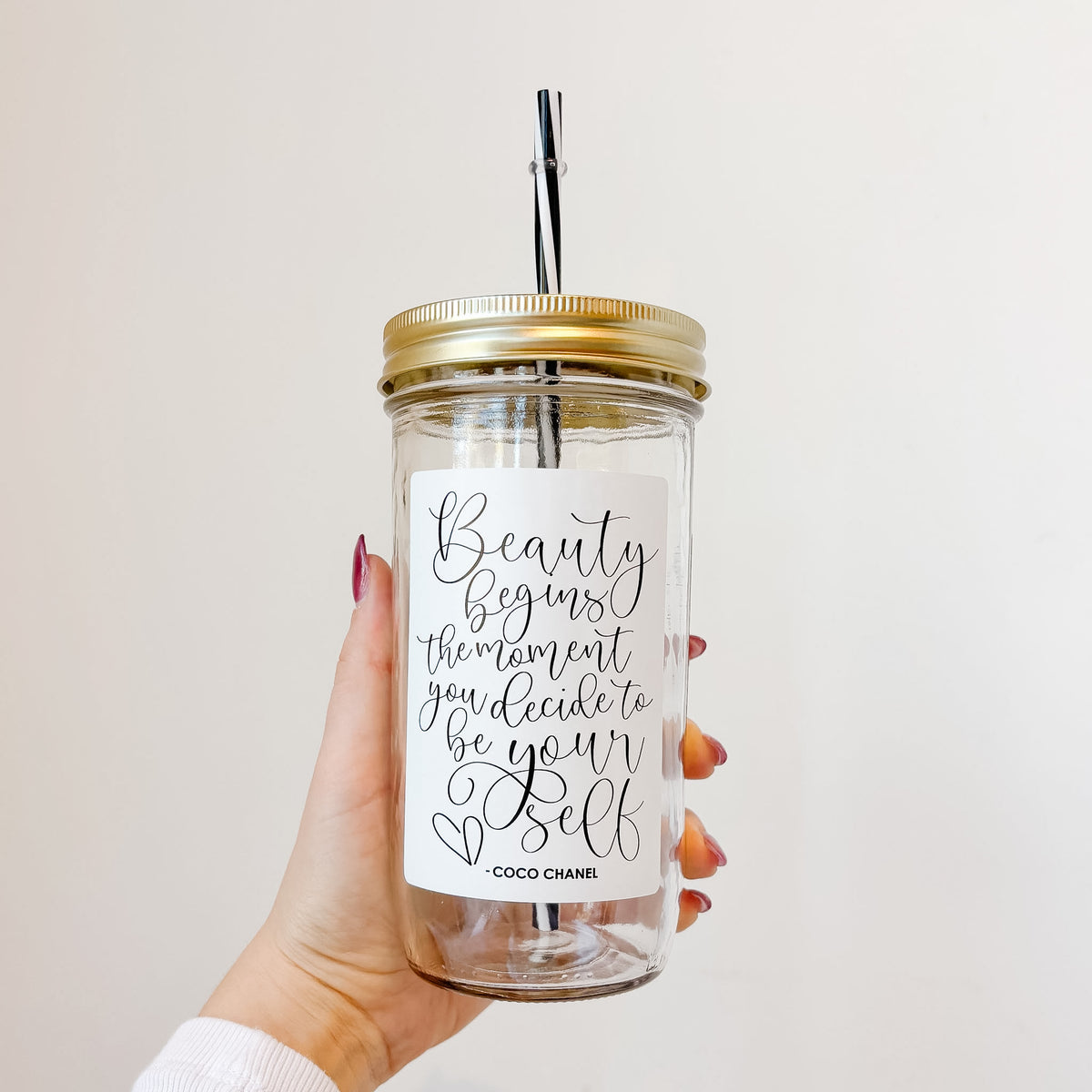 A hand holding a clear glass mason jar tumbler with a gold metal lid and black-and-white striped straw. The jar has a white design with black script text reading "Beauty begins the moment you decide to be yourself- Coco Chanel".