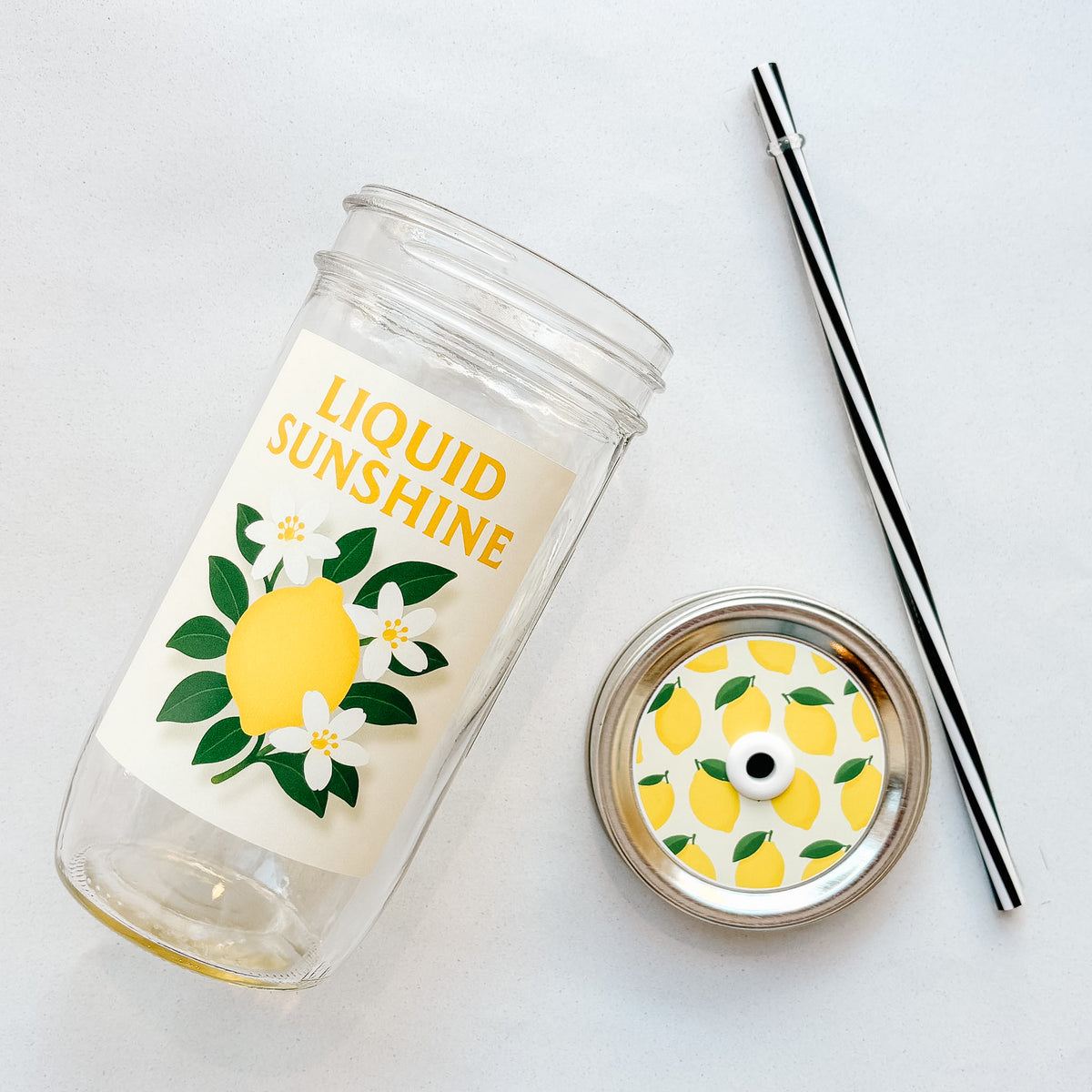 A clear glass tumbler with a pale yellow design reading 'LIQUID SUNSHINE' and a yellow lemon illustration, shown with a matching lemon-patterned lid and a black-and-white striped straw on a light background.