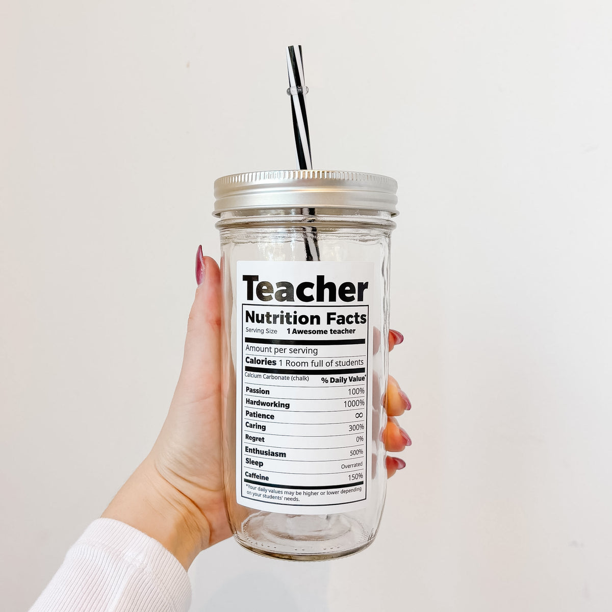 A hand holds a clear glass mason jar tumbler with a silver lid and black-and-white striped straw. The front of the glass features a white label styled like a nutrition facts panel with the word 'Teacher' at the top.