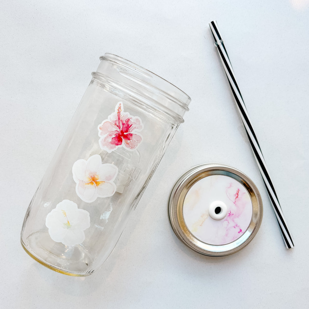 A clear glass tumbler with a pink, peach, and white hibiscus flower, shown with its matching pink and yellow watercolor lid and a black-and-white striped straw on a light background.